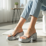 Person wearing transparent platform sandals in a bright room with a white chair and plant.